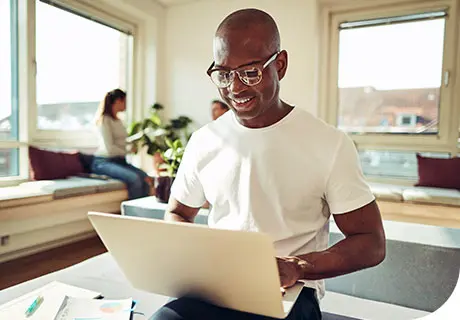 A man in a white t-shirt and glasses sits on a table edge with his laptop researching about the levels of zinc in denture adhesive. 