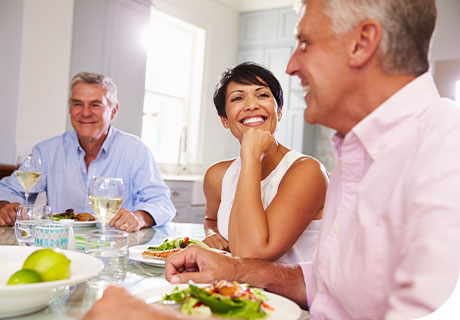 Two men and a woman in their fifties are sitting around the table chatting. The woman has a confident smile as she has no concerns about smiling with dentures.