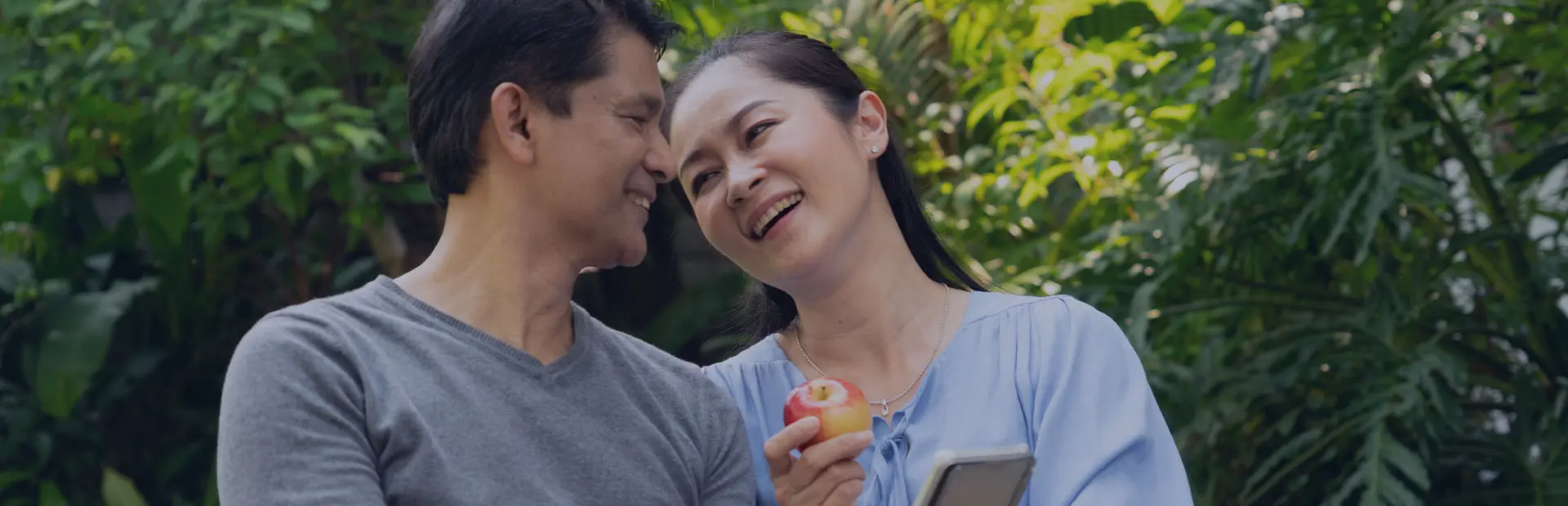 A couple in their fourties are sitting in a park enjoying a meal together, the woman shows the man her phone with tips on how to eat healthily with dentures.