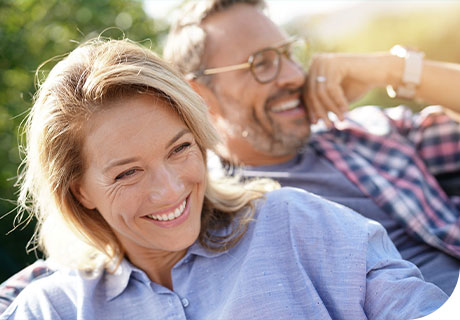 A couple are outdoors smiling in the sun, as they are getting used to wearing new dentures. 