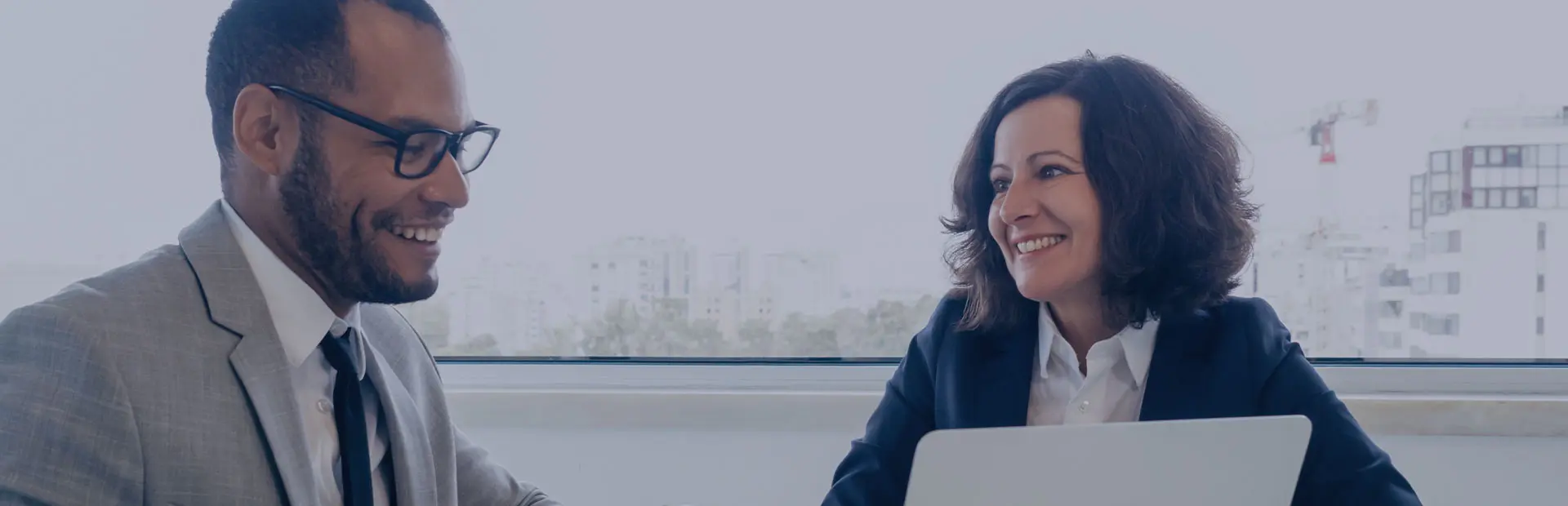 In a white office with a large window, two colleagues sit and talk confidently over a presentation given on a white laptop, one of them has dentures and wears them with confidence. 