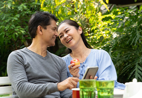 A couple in their forties are sitting in a park enjoying a meal together, the woman shows the man her phone with tips on how to eat healthily with dentures. 