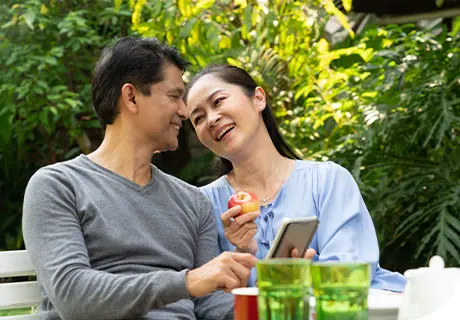 A couple in their forties are sitting in a park enjoying a meal together, the woman shows the man her phone with tips on how to eat healthily with dentures. 