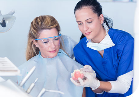 A woman is sitting in the dentist chair with her dentist holding a set of dentures in her hands as she helps answer the frequently asked questions she has about denture size and gagging. 