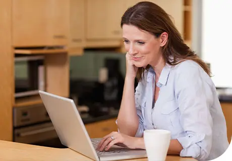 A woman in her 40s is researching about how her life can be improved with Fixodent on her laptop as she sits in a wood-lined kitchen. 