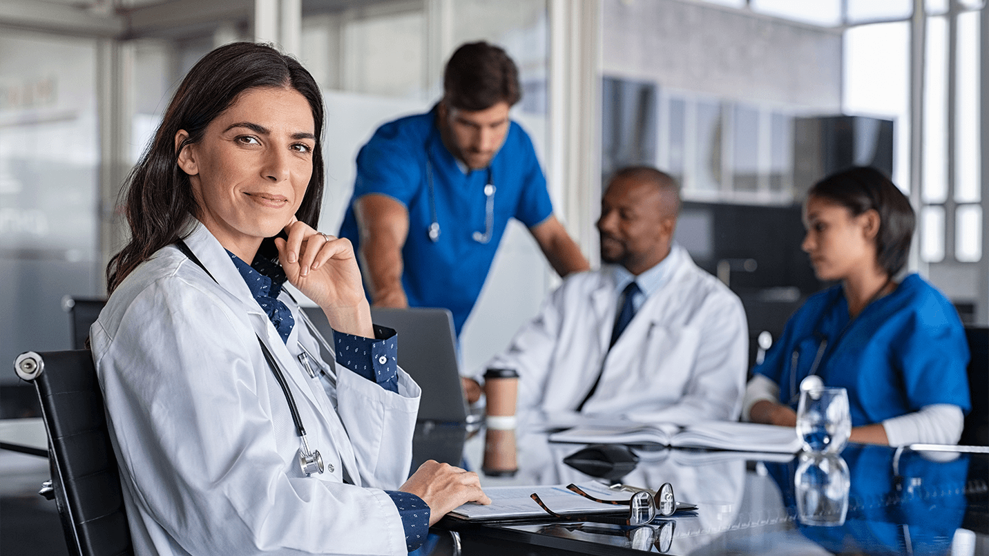 female physician sitting in a chair smiling at the camera with three other physicians in the background working.