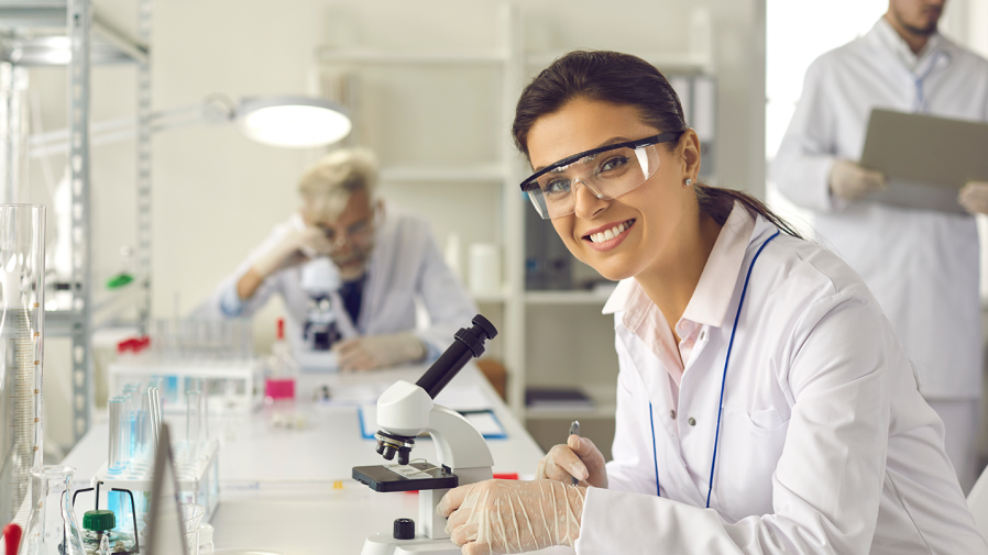 lab technician smiling at the camera while using a microscope