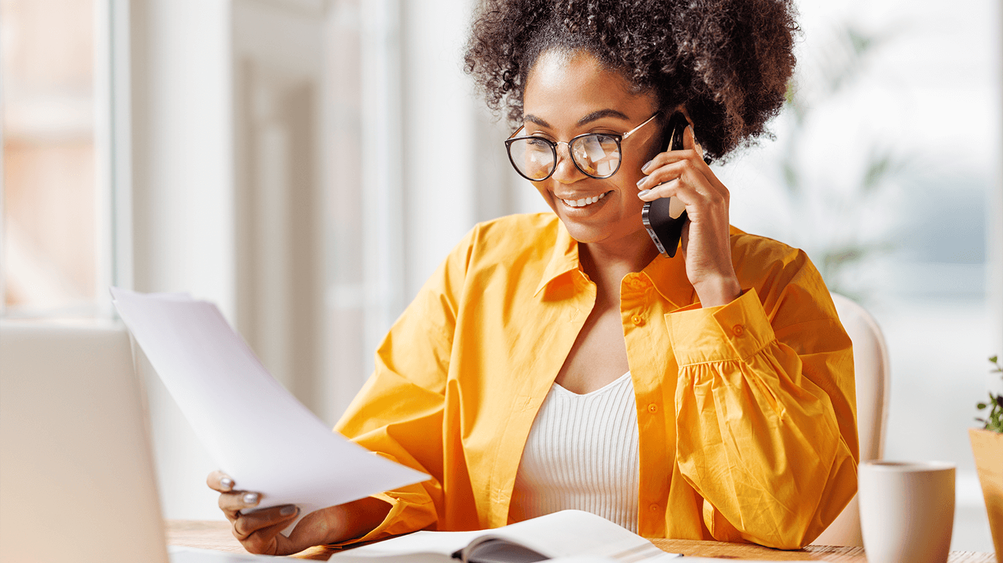 Smiling, young black female wearing glasses while sitting at desk, talking on the phone and reviewing paperwork