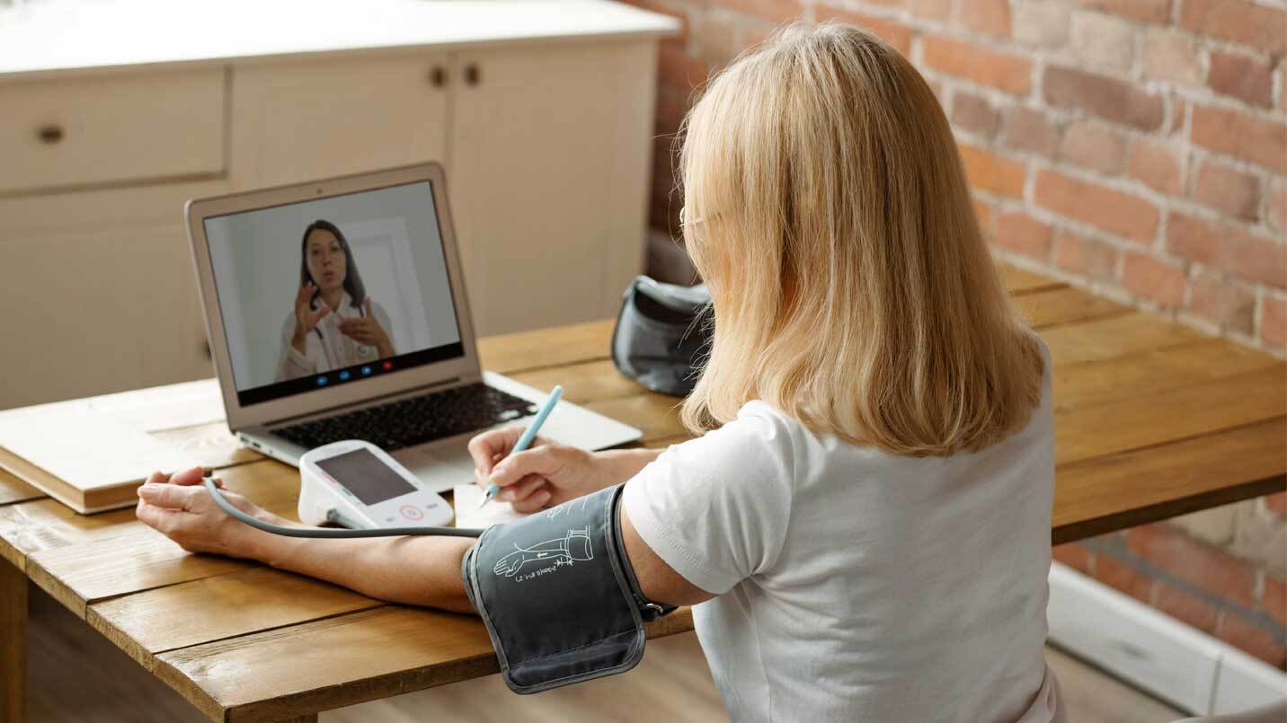 Woman using an at-home blood pressure cuff while video chatting with a physician on a laptop