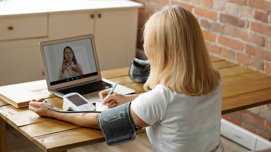 Woman using an at-home blood pressure cuff while video chatting with a physician on a laptop