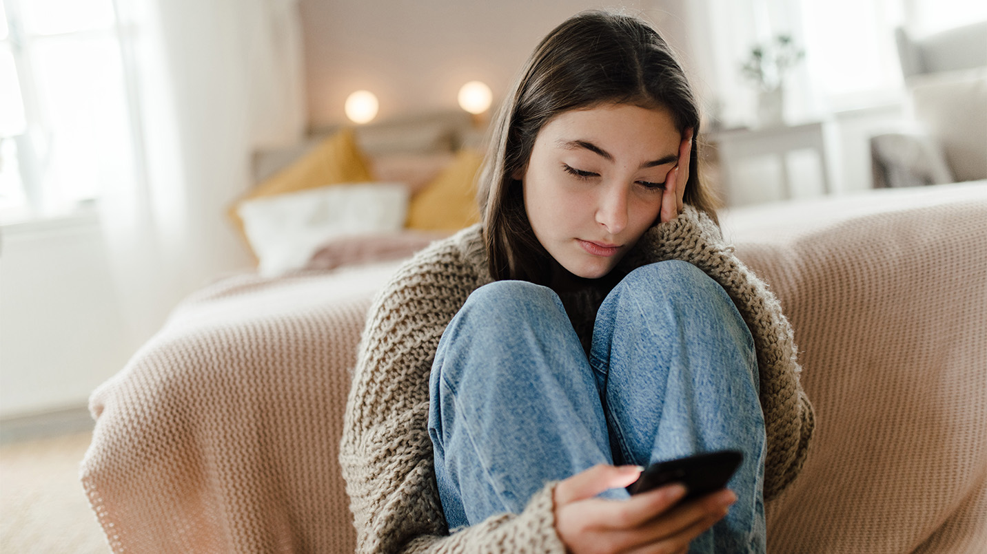teenage girl on her phone and sitting at the end of her bed with a sad expression