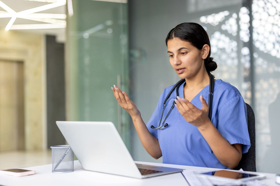 Woman doctor sitting at a desk in front of a laptop; she is conducting a telehealth consultation. 