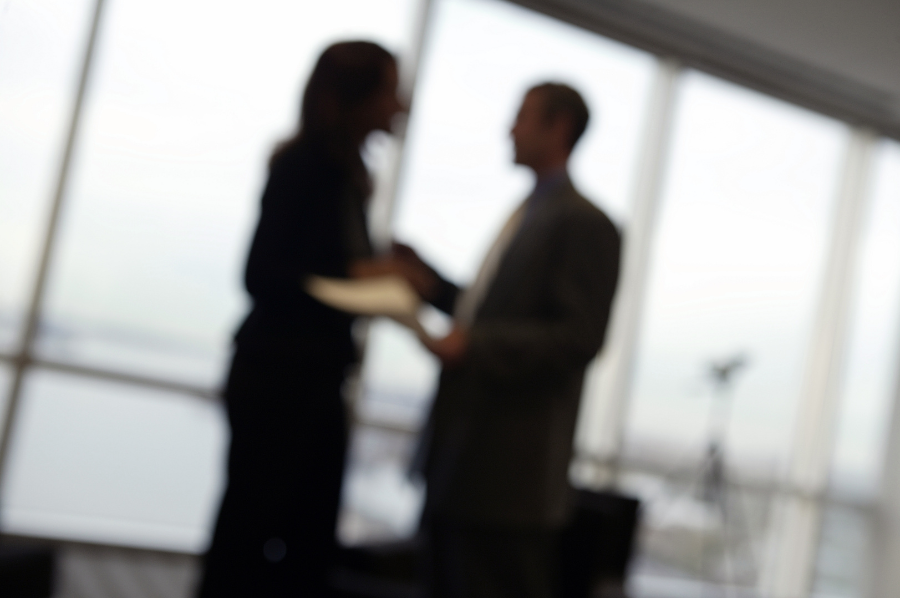 An image of a man and woman standing and speaking in a corporate office. They are in the background and out of focus, which makes the nature of their interaction unclear. They may be having an argument or a tense discussion.