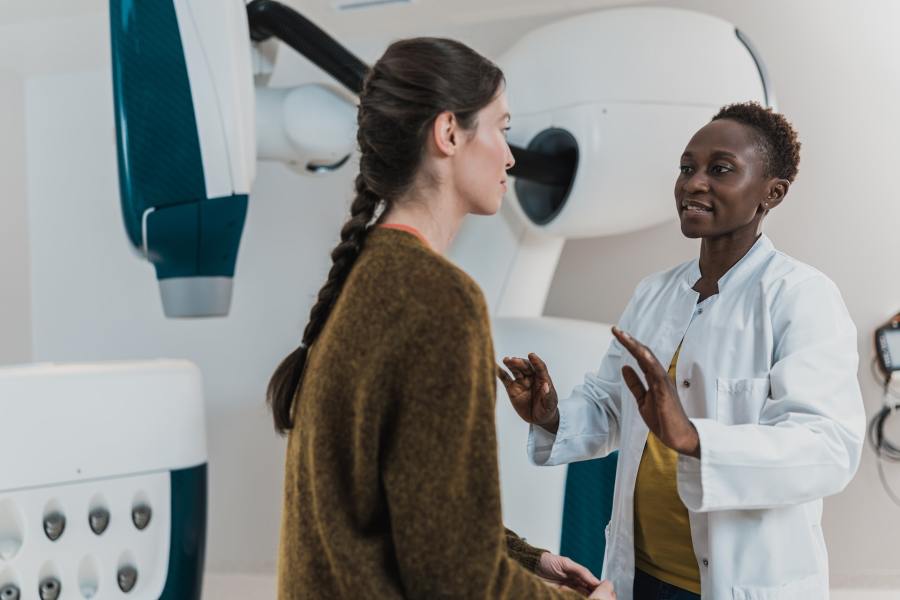 Doctor in a white lab coat is speaking to a patient seated in front of her. In the background, a radiotherapy treatment machine can be seen.