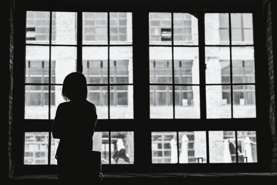 A black and white photograph of a woman with her back to the camera, looking out of a large window at the street and building outside.