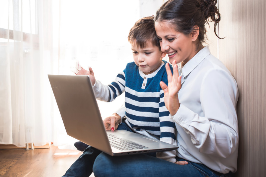 A mother with a toddler son in her lap are looking into a laptop screen and waving.
