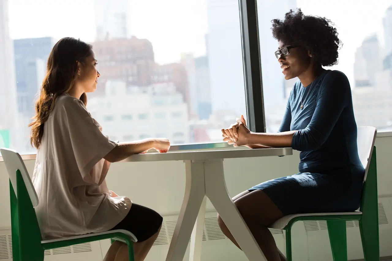 Two women sitting at a table in an office or workspace having a conversation.