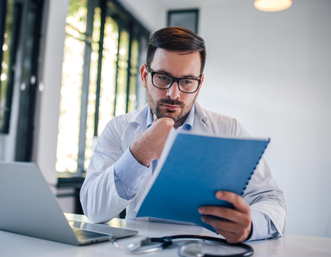 healthcare professional reviews a medical document while sitting at a desk with a laptop and stethoscope in a bright office.