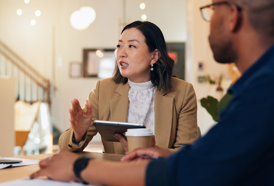 Two people sit at a table, a man looking away from the camera is in the foreground and a lady is facing the camera. She holds an ipad in her hand and there is a takeaway coffee cup on the table. Both people are wearing professional business attire. 