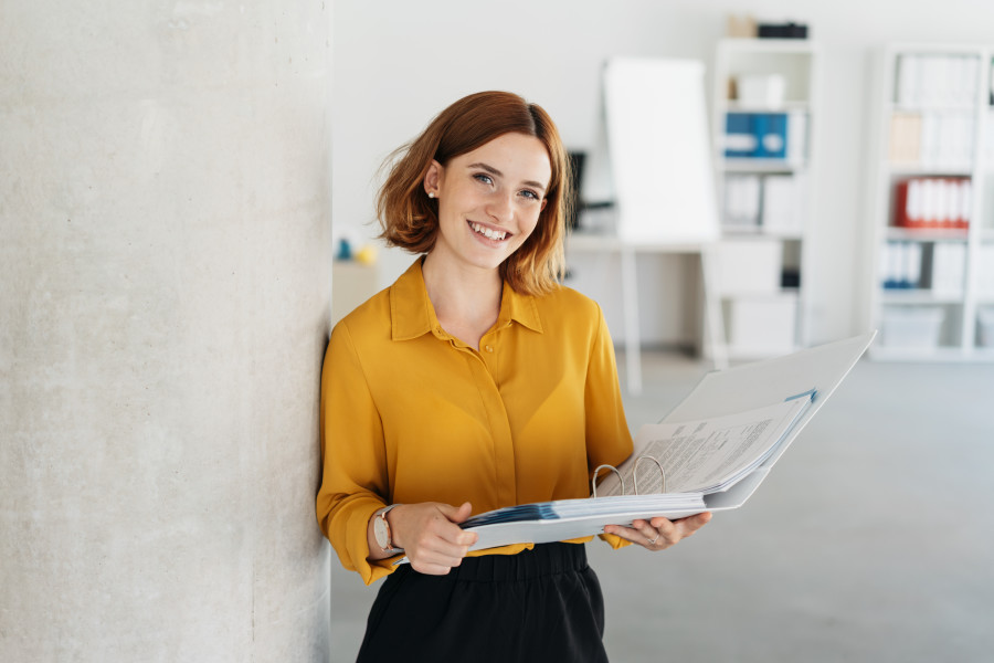 A woman in a yellow shirt is standing with her back against a white wall in an office and holding an open document folder. She is smiling at the camera in a friendly fashion.