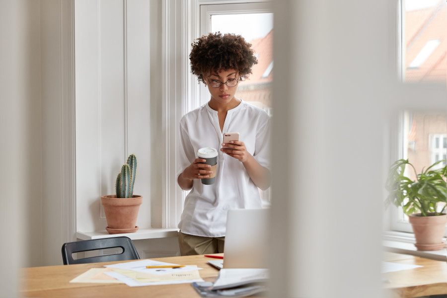 Woman standing next to a window, holding a coffee in one hand and her mobile phone with the other. She is looking at her phone.