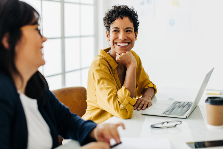 Two women sitting in an office, speaking and laughing.