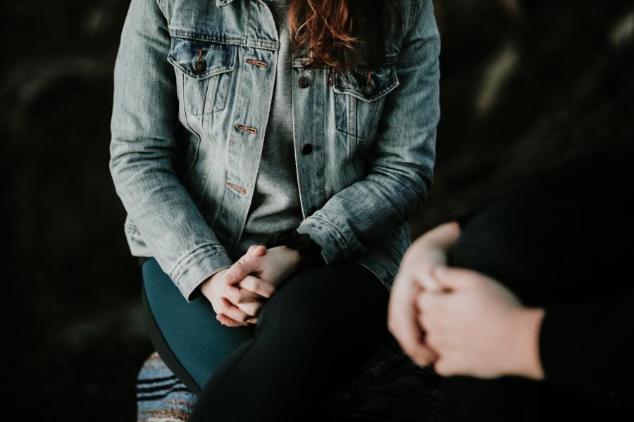 Woman in a denim jacket sitting with her hands clasped. In the foreground is a second figure in black; only their clasped hands are in shot, implying that a conversation is taking place between the two people.