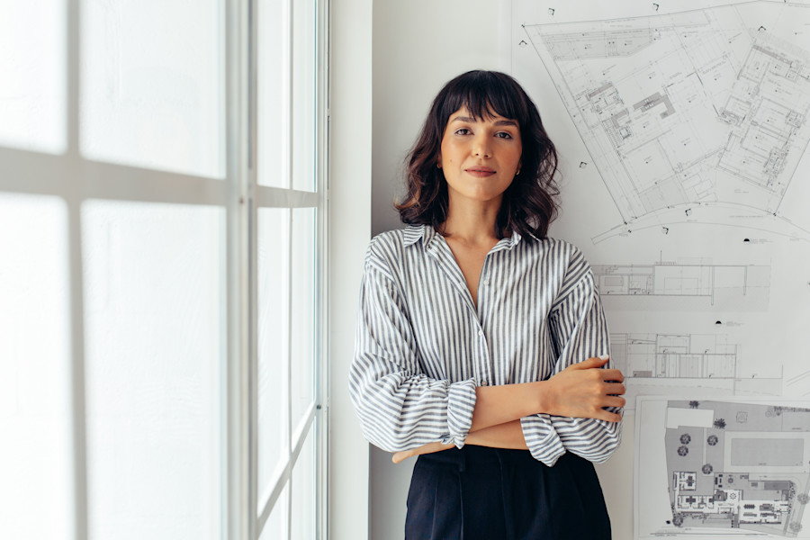Female architect standing in front of a wall of architecture drawings and plans.