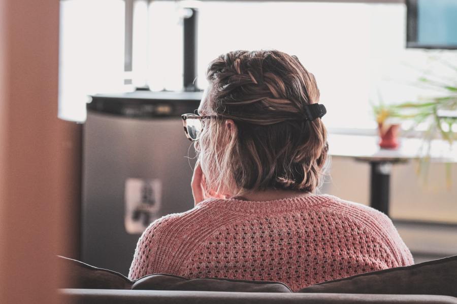 A close-up shot of the back of a woman's head. She is sitting in a waiting area, office or consulting room. She is wearing a pink-red jumper and glasses, and has short hair in a half-ponytail.