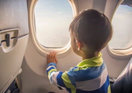 child looking out plane window at the clouds. He is wearing a yellow, blue and white stripped polo and has one hand on the bottom of the window 