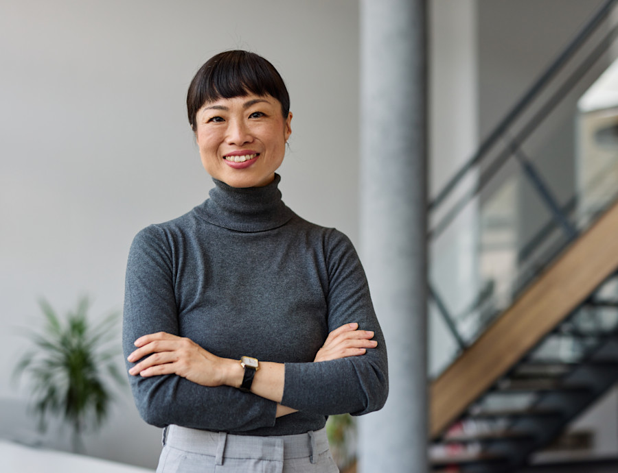 Woman standing with arms crossed, facing forward and smiling. She is an office or contemporary work or studio environment. 