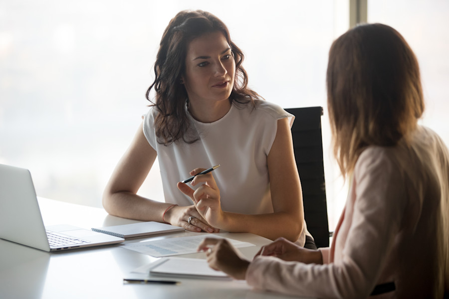 Two women in an office sitting at a desk having a conversation over some documents.