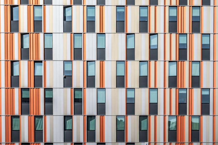 An abstract image of a building facade. The multi-storey building has narrow vertical windows and vertical cladding panels in white, peach and orange tones.