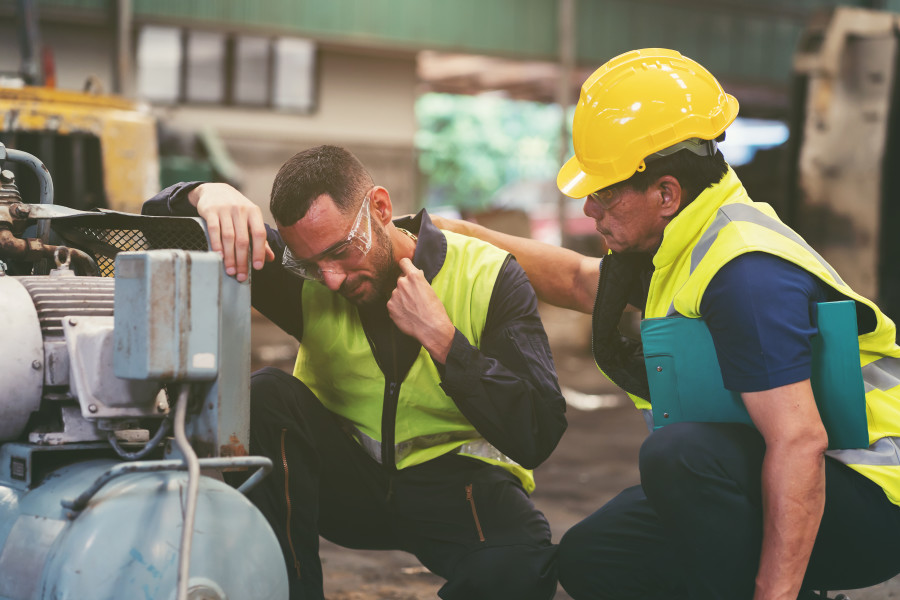 Injured factory worker in protective glasses and high-vis yellow vest is crouching down next to a piece of machinery holding his neck in pain, while his colleague or supervisor comforts him. 