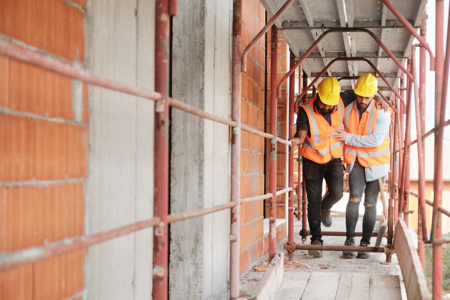 Two construction workers on a worksite. One is hobbling with an injured foot or leg, and the other is supporting him and helping him to walk.