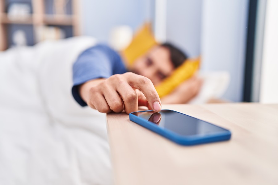 Man lying in bed, reaching his hand out to turn off his alarm or refuse a call coming through to the mobile phone lying on the sidetable next to him. 