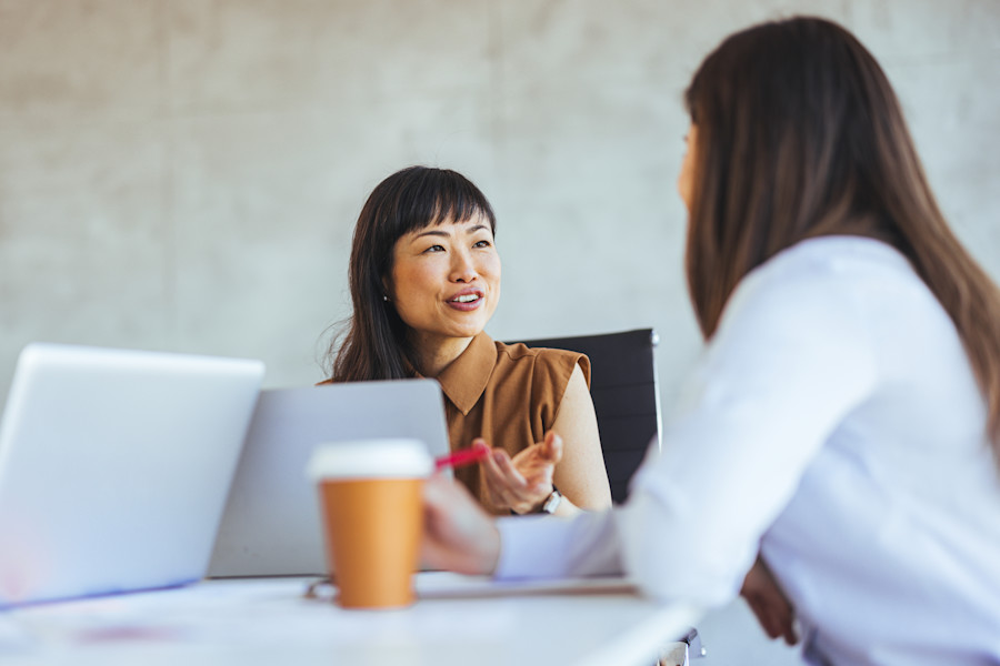 Smiling woman speaking to another woman while sitting at a desk or table drinking coffee.
