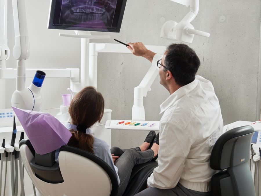 A woman is sitting in a dentist chair while her dentist sits next to her explaining an x-ray displayed in front of them.