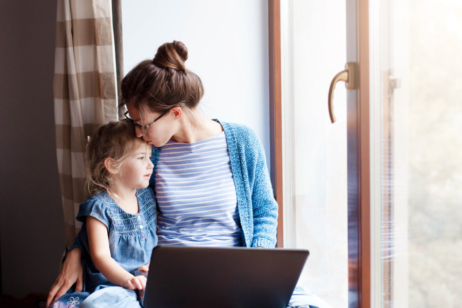 Mother and daughter sitting next to a window. They have a laptop open in front of them and the mother is kissing the daughter on the forehead.