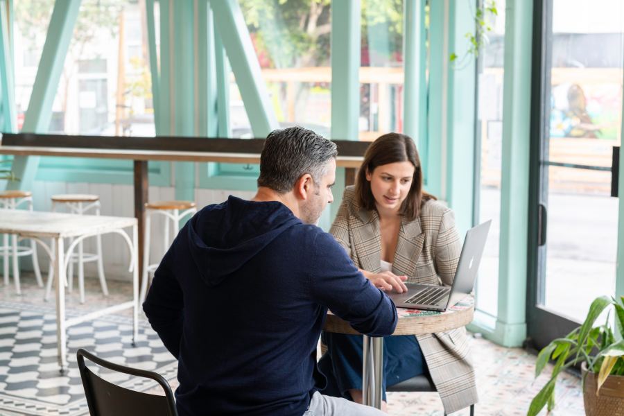 A man and woman are sitting at a table in a cafe or co-working space discussing something over an open laptop.