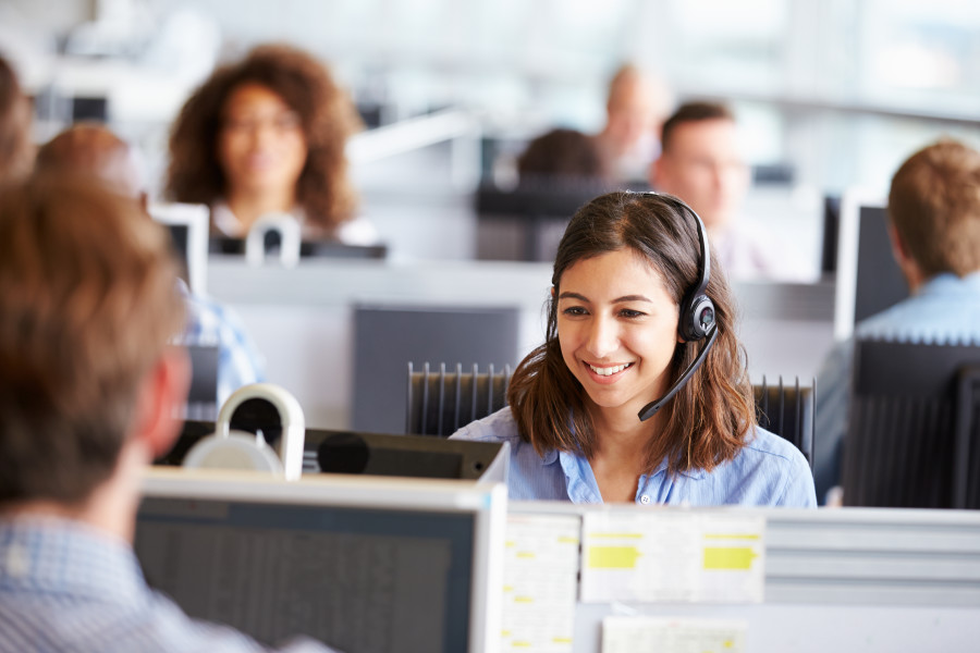 A woman working in a call centre. She is sitting at her desk wearing a headset and smiling.
