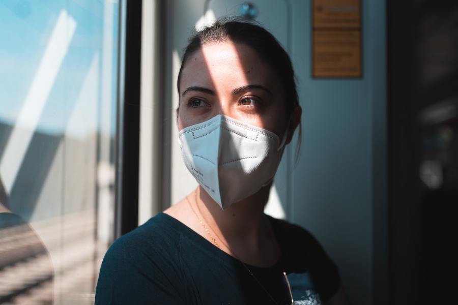 A surgeon in a surgical mask is standing near a window looking to the side. The light from the window casts striped shadows across her face.