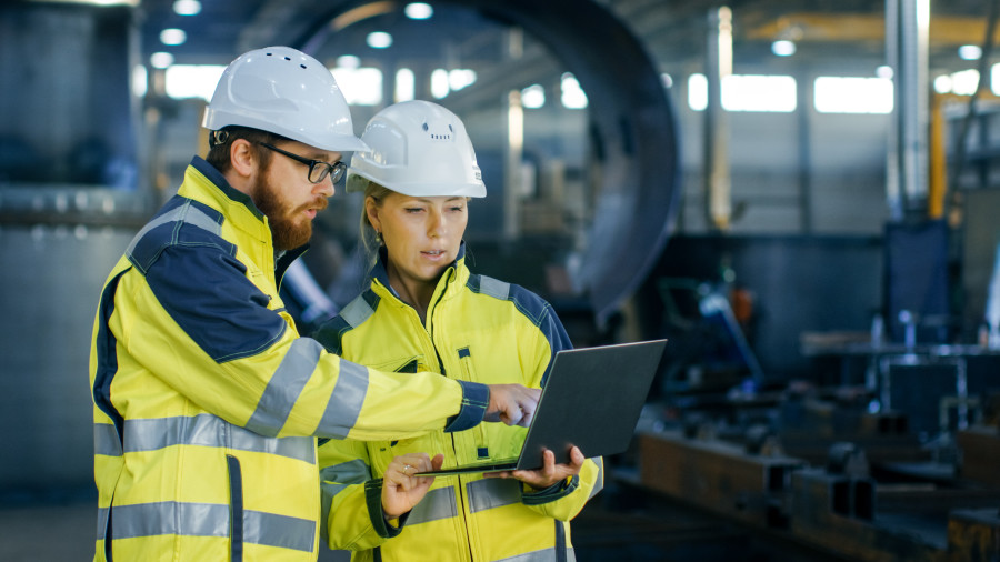 Male engineer and female engineer dressed in high-vis jackets and hard hats are discussing while looking at a laptop they are holding open between them.