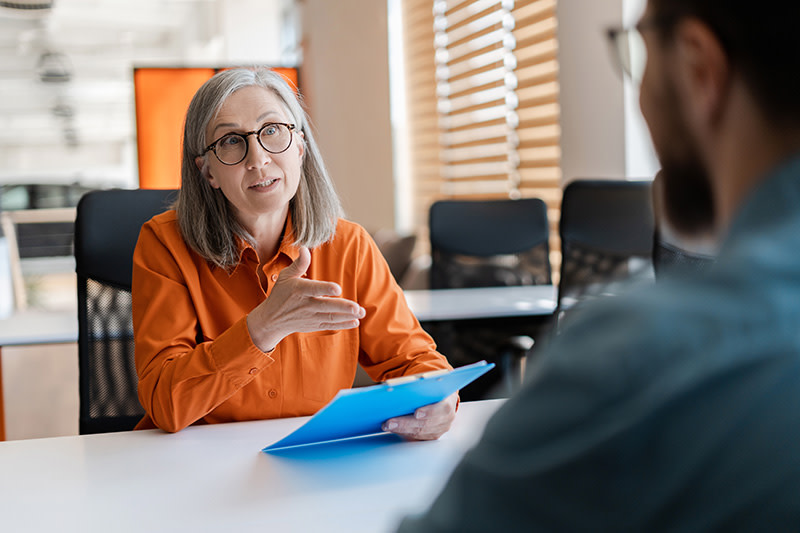 Senior manager or HR professional sitting at a desk holding a document. She is speaking and gesticulating to a man seated in front of her.