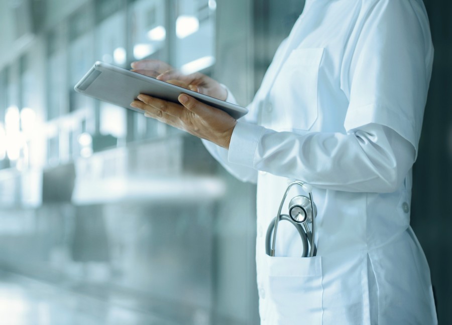 Image shows the torso of a doctor or medical professional standing in the corridor of a hospital or healthcare facility. They are wearing a white medical jacket and shirt and have a stethoscope peeking out of their hip pocket. They are holding an iPad or digital tablet in their hands.