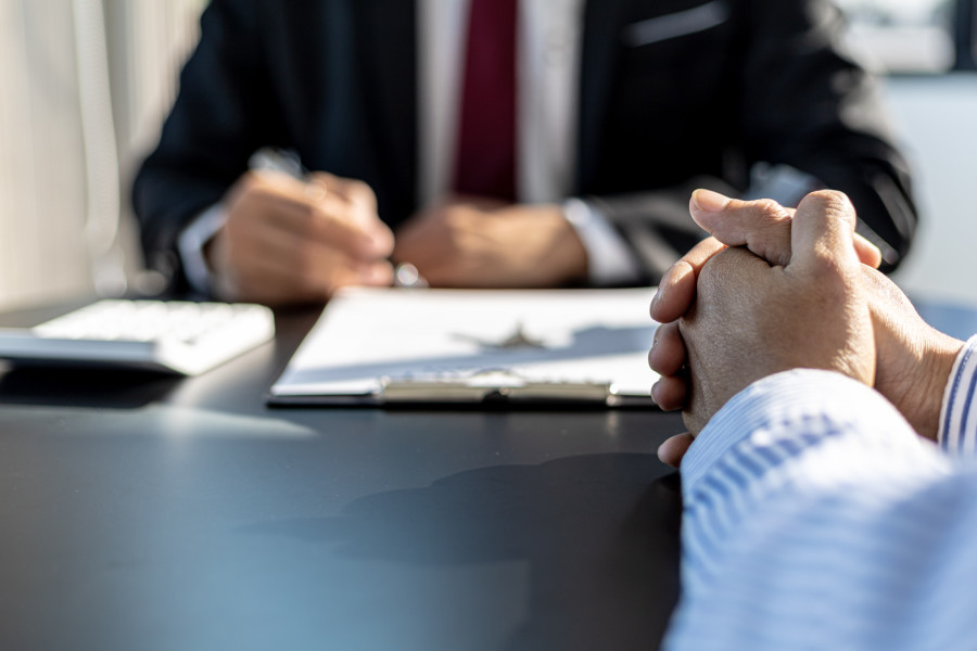 Legal or financial advisor dressed in a suit sitting at desk in front of a client, who has their hands clasped together.