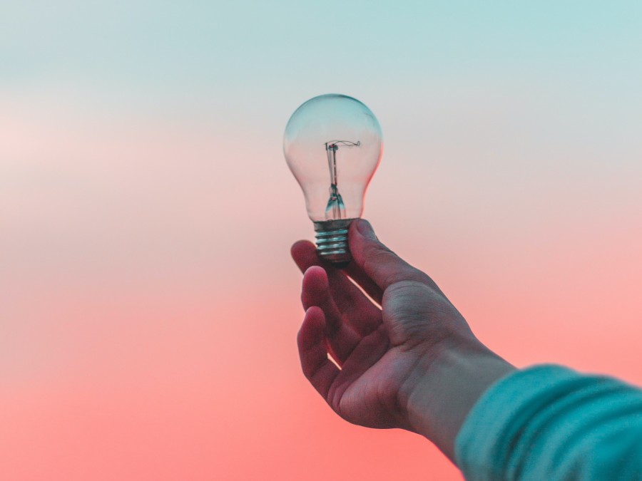 Close-up of an arm holding out a lightbulb in front of a pink and blue sunset background.