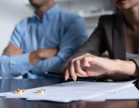 man crossing his arms, sitting at a table with a woman who is signing what appears to be a contract. Their wedding rings are on the table. 