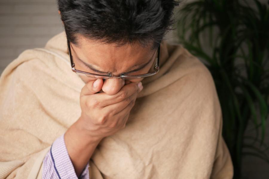 A man sneezing or coughing and holding a tissue to his face due to a cold, flu or other infection. He is wrapped in a blanket for warmth.