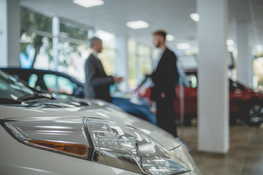 A close shot of a white car in a dealership showroom, with a salesperson and customer pictured in the background.